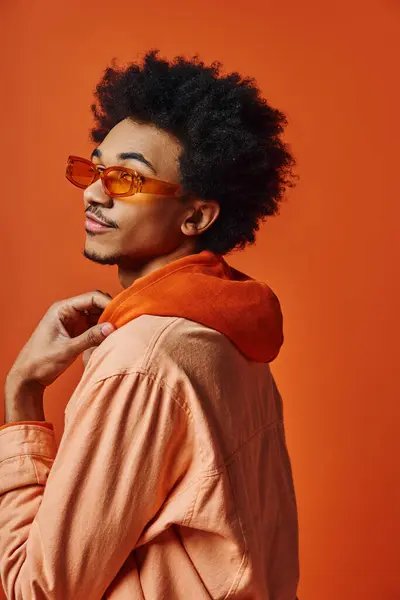 Um jovem afro-americano elegante com cabelo encaracolado usa óculos de sol e um traje na moda em um fundo laranja. — Fotografia de Stock