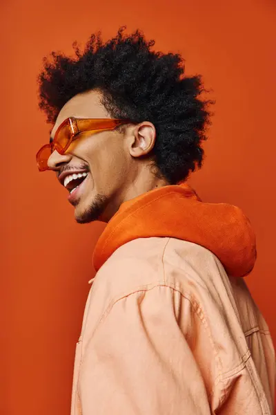 A stylish young African American man with a curly afro wearing sunglasses and a jacket, expressing emotions on an orange background. — Stock Photo