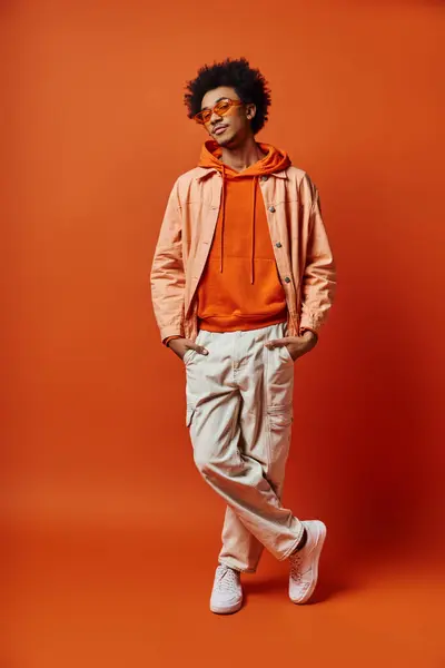 Trendiger afroamerikanischer Mann mit lockigem Haar, trägt Sonnenbrille, posiert emotional in leuchtend orangefarbenem Kapuzenpulli und khakifarbener Hose. — Stockfoto