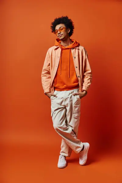 Trendy African American man with curly hair, wearing sunglasses, posing emotionally in bright orange hoodie and khaki pants. — Stock Photo