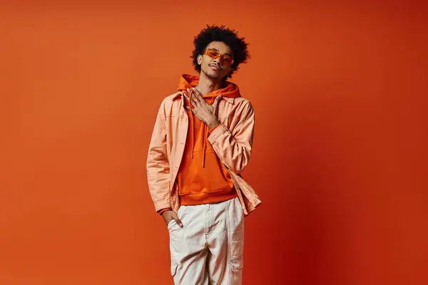 A trendy, curly African American man poses in an orange jacket and white pants against a bold orange backdrop, exuding confidence and style. — Stock Photo