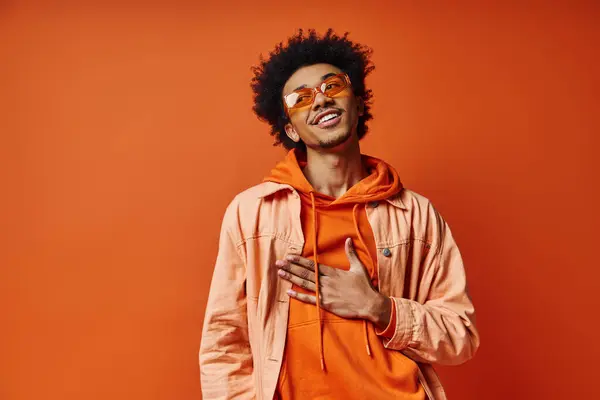Ein lockiger afroamerikanischer Mann strahlt Emotionen in trendigem orangefarbenem Kapuzenpulli und Sonnenbrille vor leuchtend orangefarbenem Hintergrund aus. — Stockfoto