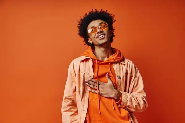 Un homme afro-américain bouclé dégage une émotion dans un sweat à capuche orange tendance et des lunettes de soleil sur fond orange vif. — Photo de stock