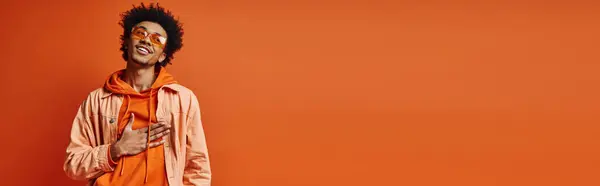Un jeune homme afro-américain élégant avec des cheveux bouclés et des lunettes de soleil portant une tenue orange et blanche, exsudant l'émotion. — Photo de stock