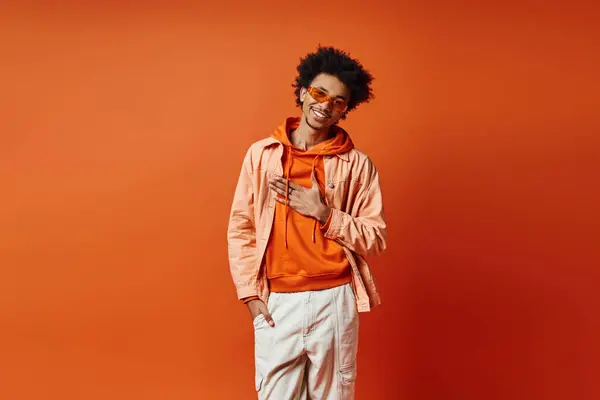 Stylish young African American man with curly hair wearing trendy orange jacket, white pants, and sunglasses on vibrant background. — Stock Photo
