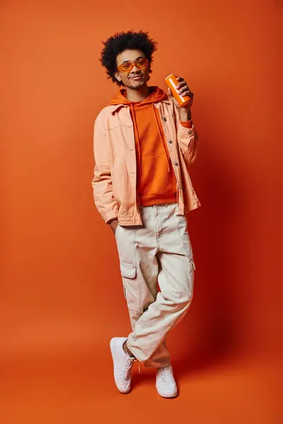 Un hombre afroamericano de moda con el pelo rizado y gafas de sol de pie con confianza sobre un fondo naranja vibrante. - foto de stock