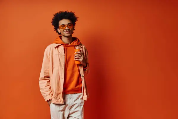 Ein angesagter afroamerikanischer Mann in orangefarbenem Kapuzenpullover und Sonnenbrille genießt einen Drink vor einem leuchtend orangen Hintergrund. — Stockfoto