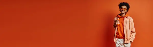 Stilvoller afroamerikanischer Mann mit lockigem Haar und Sonnenbrille, steht selbstbewusst mit den Händen in den Taschen vor einer kühnen orangefarbenen Wand. — Stockfoto