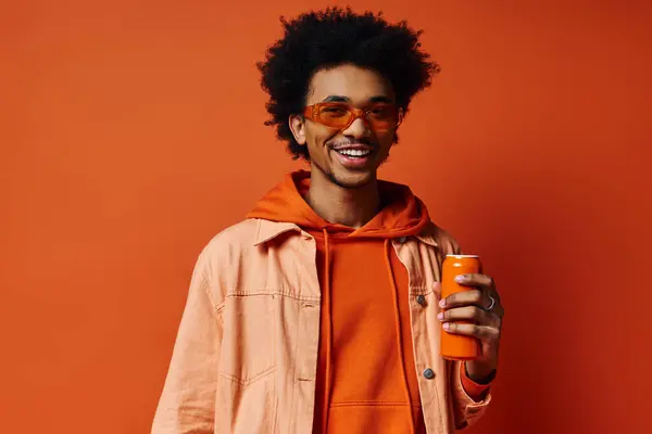 Trendiger afroamerikanischer Mann in orangefarbenem Kapuzenpulli mit einer Orangensaftdose auf leuchtend orangefarbenem Hintergrund. — Stockfoto