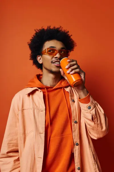 Ein angesagter afroamerikanischer Mann, lockiges Haar, orangefarbener Kapuzenpullover, Drinks aus der Dose, Sonnenbrille, emotionaler Ausdruck auf orangefarbenem Hintergrund. — Stockfoto