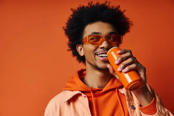 A stylish curly African American man in an orange hoodie holds an orange bottle against an orange background. — Stock Photo