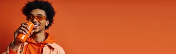 Stylish young African American man in trendy orange hoodie holding an soda can against a vibrant orange background. — Stock Photo