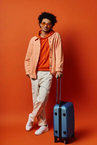 A young African American man stands next to a blue suitcase, exuding style and emotion against an orange backdrop. — Stock Photo