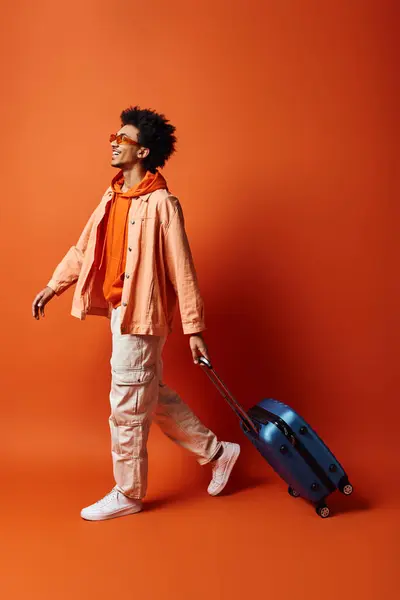 Un joven y elegante hombre afroamericano con el pelo rizado y el atuendo de moda caminando hacia adelante mientras sostiene una maleta en su mano contra un fondo naranja. - foto de stock