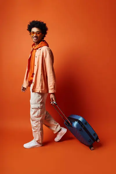 Trendy African American man with curly afro hair holding a blue suitcase against an orange background, looking emotional. — Stock Photo