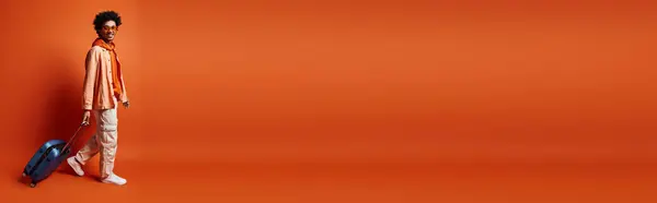 Trendy African American man with curly afro hair holding a blue luggage against an orange background, looking emotional. — Stock Photo