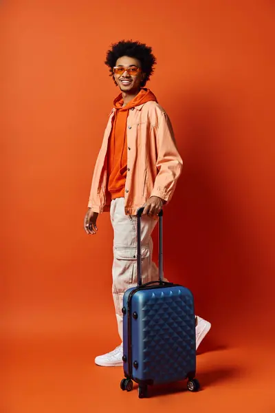 Un giovane afroamericano elegante in piedi con una valigia davanti a un muro arancione, trasudando fiducia e carisma. — Foto stock