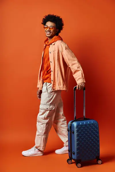 Un uomo afroamericano alla moda con una valigia si erge con fiducia su uno sfondo arancione vibrante, trasudando un senso di avventura e stile.. — Foto stock