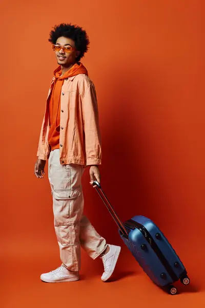 A fashionable African American man with curly hair in an orange jacket carrying a blue suitcase on an orange background. — Stock Photo