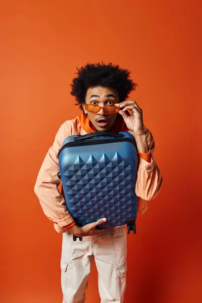 Trendy African American man with curly hair holding a stylish blue luggage on an orange background. — Stock Photo