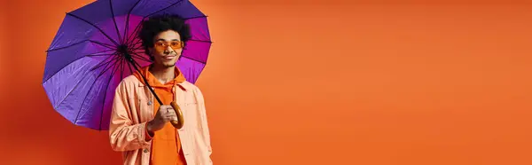 A trendy young African American man with curly hair, sunglasses, and fashionable attire holds a vibrant purple umbrella on an orange background. — Stock Photo