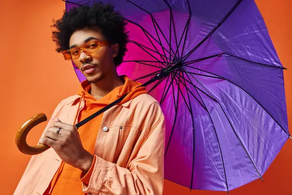 Giovane uomo afroamericano con capelli ricci e occhiali da sole che tiene un ombrello viola sopra la testa su uno sfondo arancione. — Foto stock