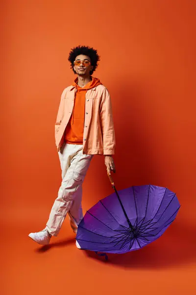 Un hombre afroamericano joven y elegante con el pelo rizado está junto a un paraguas púrpura sobre un fondo naranja. - foto de stock