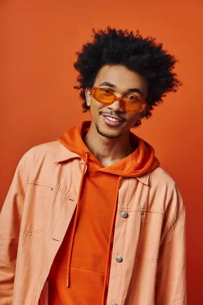 Стильный молодой афроамериканец с вьющимися волосами, одетый в оранжевую рубашку и куртку на оранжевом фоне, демонстрирующий различные эмоции. — стоковое фото
