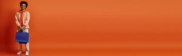 Trendy African American guy in sunglasses stands with blue bag against orange backdrop, exuding emotional energy. — Stock Photo