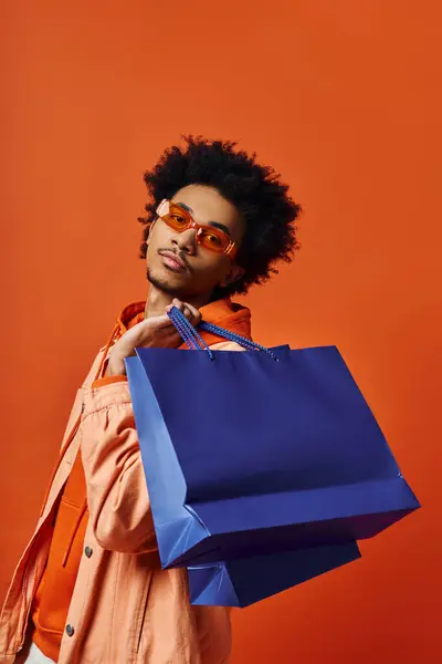 Un hombre afroamericano rizado con un atuendo moderno sostiene una bolsa de compras azul sobre un fondo naranja, con un aspecto emocional. - foto de stock