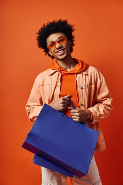 Stylish young African American man in sunglasses holding a blue shopping bag against an orange background. — Stock Photo