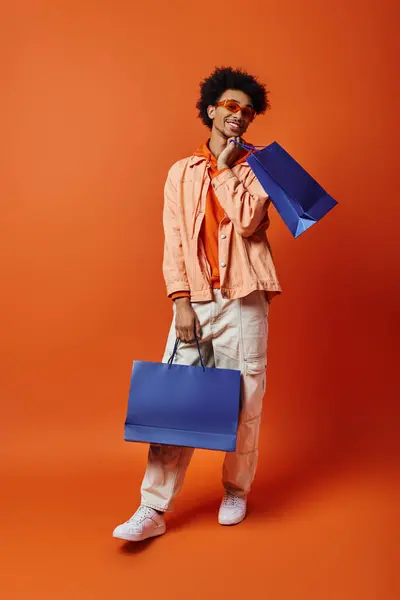 Homem afro-americano encaracolado em trajes da moda mantém um saco azul enquanto mostra um saco azul único e vibrante contra um fundo laranja. — Fotografia de Stock