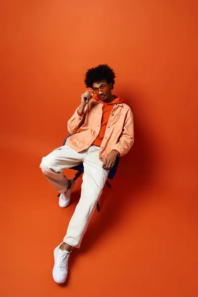 Jeune homme afro-américain à la mode en tenue tendance et lunettes de soleil assis sur un mur orange audacieux, perdu dans la pensée. — Photo de stock