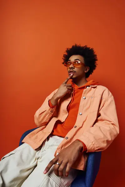 Um jovem afro-americano elegante com cabelo encaracolado sentado em uma cadeira azul, vestindo uma camisa laranja, perdido em pensamento. — Fotografia de Stock