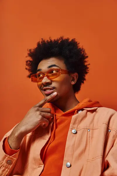 A trendy young African-American man with curly hair wearing an orange shirt and sunglasses poses against an orange background. — Stock Photo