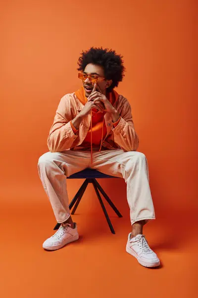 Ein stilvoller junger afroamerikanischer Mann mit lockigem Haar und Sonnenbrille sitzt kühn auf einem Stuhl vor orangefarbenem Hintergrund.. — Stockfoto