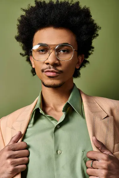 Un joven y elegante hombre afroamericano con el pelo rizado con atuendo de moda y gafas de sol sobre un fondo verde. - foto de stock