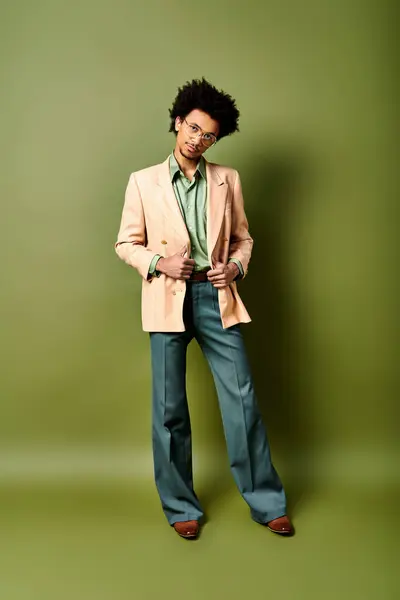 Joven hombre afroamericano con estilo en un traje y gafas de sol se coloca con confianza frente a una pared verde vibrante. - foto de stock