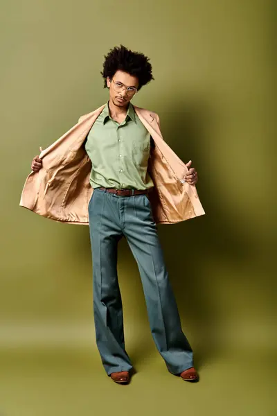 A stylish young African American man with curly hair standing confidently in front of a bold green wall while wearing trendy attire and sunglasses. — Stock Photo