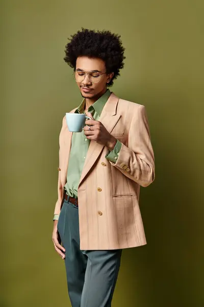 Jeune homme afro-américain élégant en costume et lunettes de soleil, tenant une tasse de café sur un fond vert vibrant. — Photo de stock