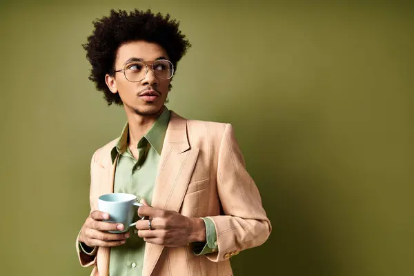 Elegante joven afroamericano en traje de moda y gafas de sol, saboreando una taza de café sobre un fondo verde. - foto de stock