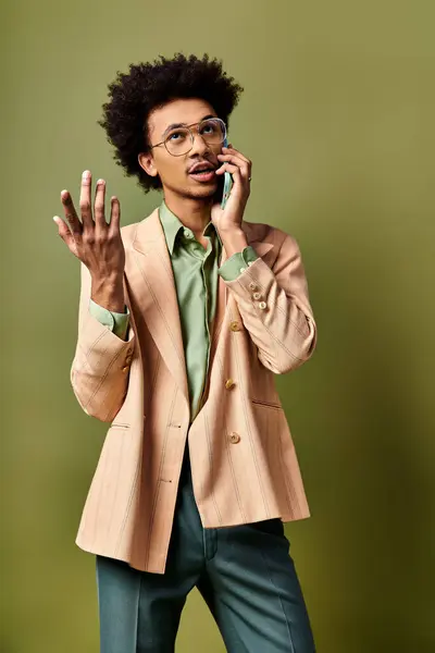 Un hombre afroamericano joven y elegante con un traje y gafas de sol hablando en un teléfono celular sobre un fondo verde vibrante. - foto de stock