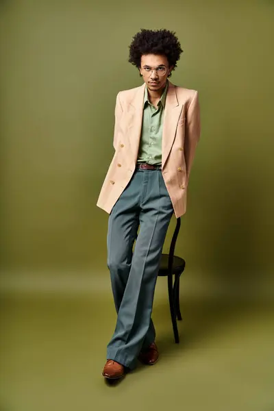 Стильний молодий афроамериканський чоловік у костюмі та сонцезахисних окулярах стоїть впевнено поруч зі стільцем на зеленому тлі. — стокове фото