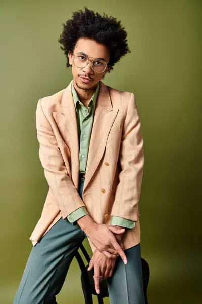 Un giovane afroamericano elegante, vestito e occhiali, si siede con fiducia su una sedia sullo sfondo verde. — Foto stock