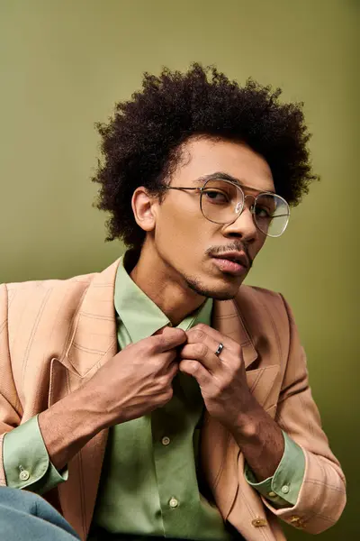 Стильний молодий афроамериканський чоловік у костюмі та окулярах ретельно застібає сорочку на яскраво-зеленому тлі. — стокове фото