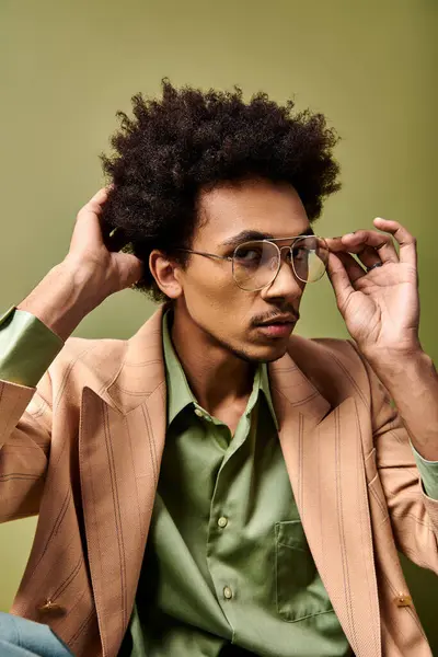 Стильний молодий афроамериканський чоловік з кучерявим волоссям, гострим костюмом та сонцезахисними окулярами, сидить на зеленому фоні. — стокове фото