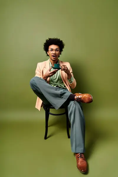 Stylish African American man with curly hair sitting on a chair, holding a cell phone, wearing trendy clothes and sunglasses, against a green background. — Stock Photo