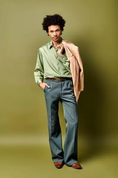 Elegante giovane uomo afro-americano con i capelli ricci indossa una camicia verde e pantaloni blu, occhiali da sole sportivi su uno sfondo verde. — Foto stock