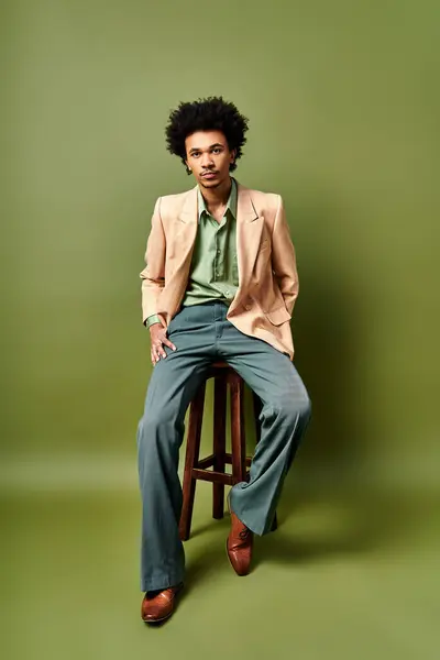 A trendily dressed young African American man with curly hair sitting atop a wooden stool against a green background. — Stock Photo
