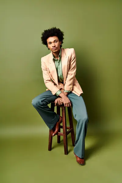 Elegante joven afroamericano de traje sentado en un taburete sobre un fondo verde. - foto de stock
